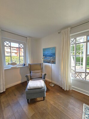 Ein Zimmer mit Holzboden, einem grauen Sessel, einem kleinen Hocker, einer Stehlampe und einem Meeresblick-Gemälde.
