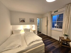 A hotel room with a bed, a nightstand, a window with curtains, and a door leading to a hallway. - Palais am Südstrand - Whg. 1 - Apartment Palais am Südstrand: Sauna, (Wyk auf Föhr)
