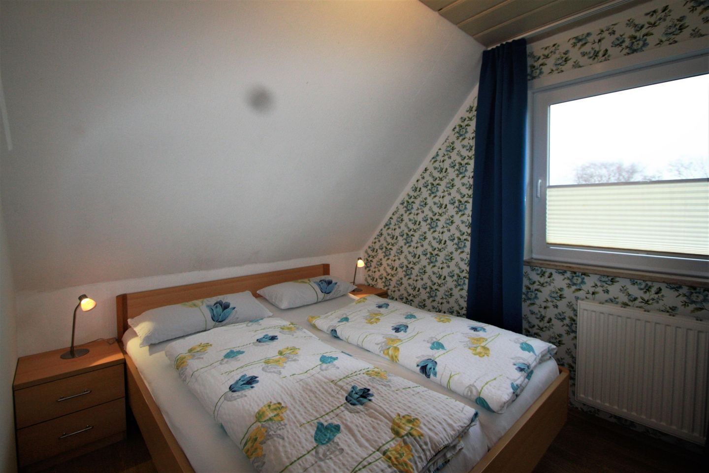 Une chambre avec un plafond en pente, un lit double avec une literie à motifs floraux, une table de chevet avec une lampe et une fenêtre donnant sur le ciel.