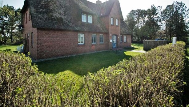 A brick house with a thatched roof, a well-maintained lawn, and a hedge in the foreground. - Bredland 2a - Whg. 5 - Holiday apartment in Nieblum: 52 m², terrace & only 200 m to the beach (Nieblum)