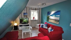 A living room with a red sofa, a white coffee table, a TV on a stand, and a painting on the wall. - Bredland 2a - Whg. 5 - Holiday apartment in Nieblum: 52 m², terrace & only 200 m to the beach (Nieblum)