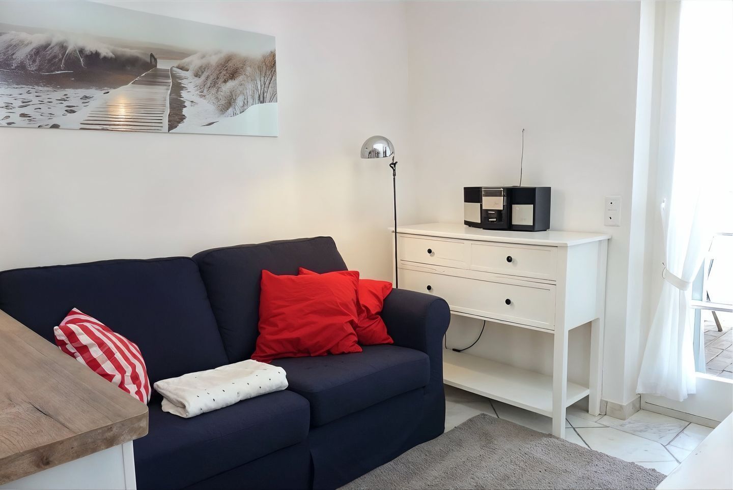 Ein Wohnzimmer mit einem dunkelblauen Sofa, roten und weißen Kissen, einem weißen Beistelltisch mit einer Lampe sowie einem Gemälde an der Wand.