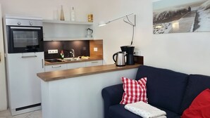 Una cocina moderna con horno integrado, cafetera y hervidor en la encimera. Hay un sofá con un cojín a cuadros rojo y blanco y una manta blanca.