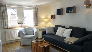 A living room with a sofa, armchair, and a small wooden table. There is a window with curtains, a radiator, and wall-mounted shelves with decorative items. - Hallig Hooge - Whg. 6 - Hbg. Hallig - Non-smoking vacation apartment on Föhr, (Wyk auf Föhr)