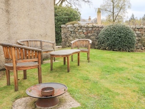 A patio with two wooden chairs and a metal table, a stone wall, and a bush in the background.