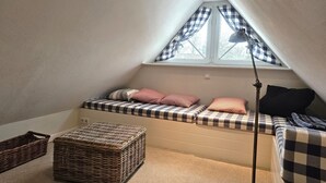 Een knus zolderkamer met schuine wanden, een raam met geruit gordijnen, een bed met geruit beddengoed, een bedlampje en twee rieten manden.