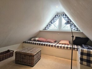 A cozy attic bedroom with a sloped ceiling, a window with checkered curtains, a bed with checkered bedding, a bedside lamp, and two wicker baskets. - Eltimjo 1 - Holiday apartment for 2 persons - Eltimjo 1 vacation apartment in Nieblum: (Nieblum)