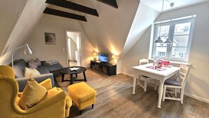 Een loft-achtige woonruimte met een eethoek, een bank, een gele fauteuil, een televisie en een raam met uitzicht over de stad.
