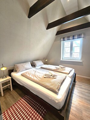 A bedroom with two beds, a window with curtains, a nightstand with a lamp, and a checkered rug. - Eltimjo 1 - Holiday apartment for 2 persons - Eltimjo 1 vacation apartment in Nieblum: (Nieblum)