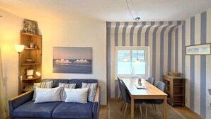 Una sala de estar con un sofá, una mesa de comedor con sillas, una estantería, un ventanal con cortinas a rayas y un cuadro de barcos en la pared.