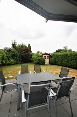 An outdoor patio with a table and chairs, a small shed, and a well-maintained hedge in the background. - Boldixumer Str. 35 - Ground floor - Holiday apartment in Wyk auf Föhr: Garden, terrace & (Wyk auf Föhr)