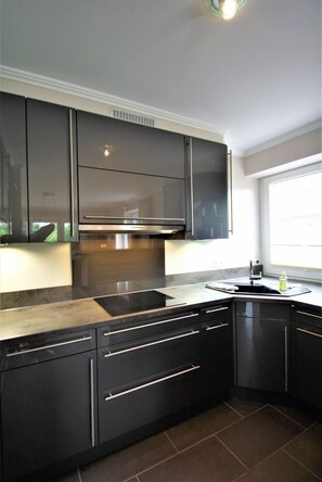 A modern kitchen with dark cabinetry, a stainless steel range hood, and a window providing natural light. - Boldixumer Str. 35 - Ground floor - Holiday apartment in Wyk auf Föhr: Garden, terrace & (Wyk auf Föhr)
