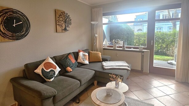 Una sala de estar moderna con un sofá modular, una mesa de centro redonda, un reloj de pared y una ventana con vistas a zonas verdes.
