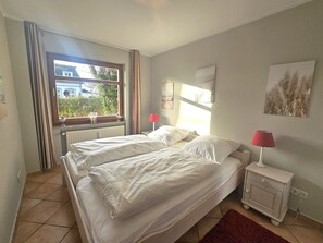 A bedroom with two beds, a window with curtains, a bedside table with a lamp, and artwork on the wall. - Schatzinsel, Whg. 1 - Holiday apartment Schatzinsel Whg. 1 on Föhr - Terrace & (Wyk auf Föhr)