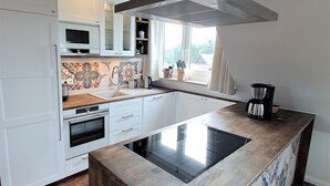 A modern kitchen with white cabinetry, a central island, a stainless steel range hood, and a window with a view. - Seestern - Holiday apartment Seestern in Wyk on Föhr - 80 m², terrace & garden (Wyk auf Föhr)