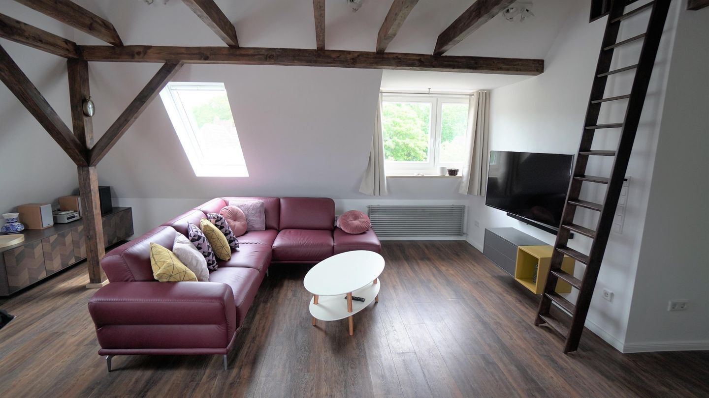 A living room with a sectional sofa, a white coffee table, wooden beams, a TV, and a window with a view of greenery.