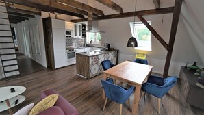 A modern kitchen with a central island, wooden beams, a dining area with blue chairs, and a staircase leading to another room. - Seestern - Holiday apartment Seestern in Wyk on Föhr - 80 m², terrace & garden (Wyk auf Föhr)