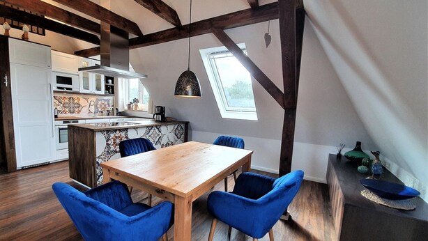 A modern kitchen with a wooden table and blue chairs, a dining area with a view of the outdoors, and a small side table with decorative items. - Seestern - Holiday apartment Seestern in Wyk on Föhr - 80 m², terrace & garden (Wyk auf Föhr)