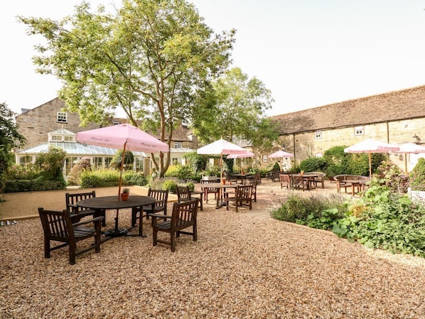 An outdoor patio with tables and chairs, surrounded by greenery and a building in the background.