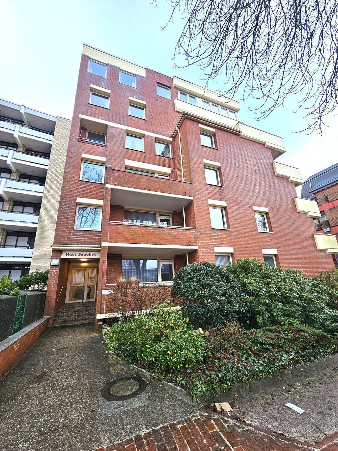 A multi-story residential building with a brick facade, multiple balconies, and a small front garden.