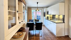 Et køkken med hvide skabe, et spiseområde med to stole, et træbord og en hængende lampe.