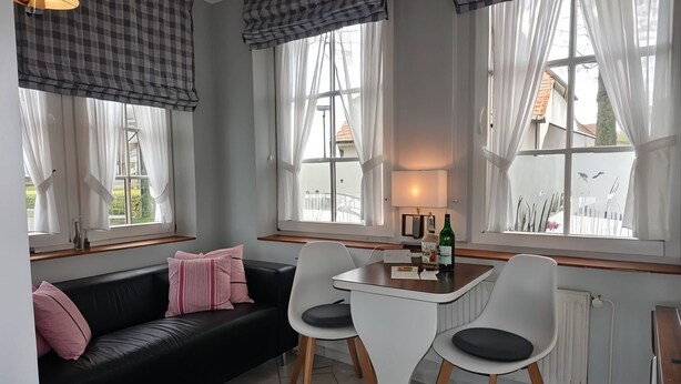 Een kamer met een zwarte bank, twee witte stoelen, een klein tafeltje en grote ramen met gordijnen.