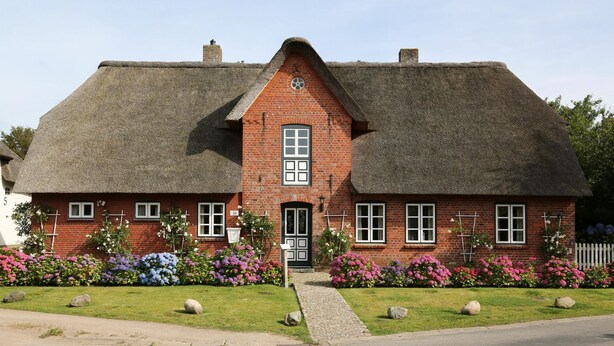 A traditional brick house with a thatched roof, a central door, and a small front garden with colorful flowers. - Ilskes Hus - Detached thatched house with garden, terrace & e-charging station (Oevenum)