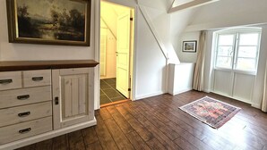 A room with a wooden floor, a cabinet with drawers, a painting on the wall, a door leading to another room, and a window with curtains. - Ilskes Hus - Detached thatched house with garden, terrace & e-charging station (Oevenum)