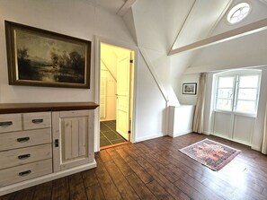 A room with a wooden floor, a cabinet with drawers, a painting on the wall, a door leading to another room, and a window with curtains. - Ilskes Hus - Detached thatched house with garden, terrace & e-charging station (Oevenum)