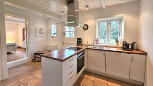 A modern kitchen with white cabinetry, a central island, a window with a view, and a built-in stove. - Ilskes Hus - Detached thatched house with garden, terrace & e-charging station (Oevenum)