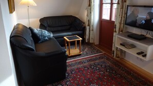 Una sala de estar con un sofá de cuero negro, una mesita de madera, un televisor sobre un mueble blanco y una alfombra estampada.