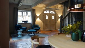A living room with a wooden table, two blue armchairs, a small white side table, a potted plant, and a staircase leading to an upper level. - Reethus main house on the ground floor/ first floor - Reethus in Borgsum: 121 m², sauna, fireplace & garden, (Borgsum)
