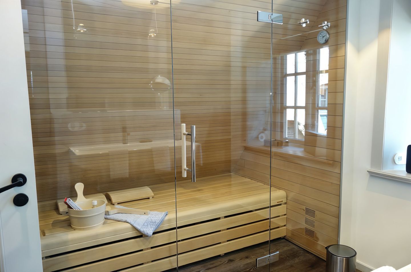 A sauna with a glass enclosure, wooden benches, a towel, and a small trash bin.
