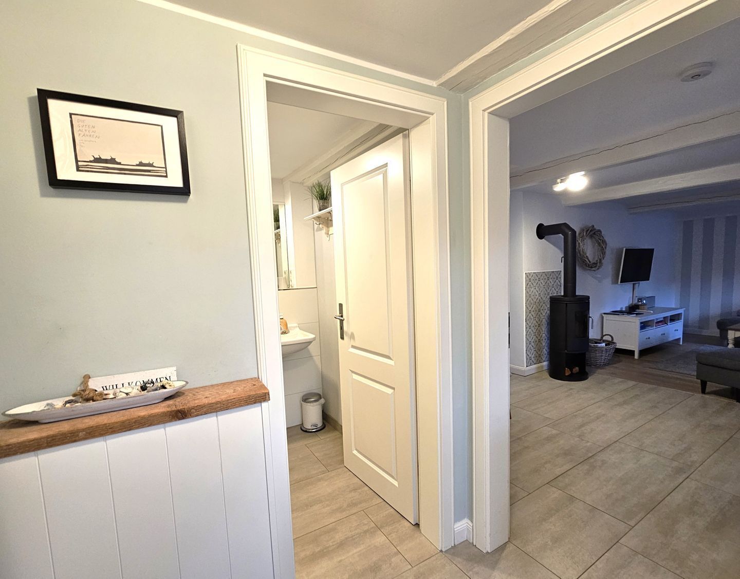 Ein Flur, der zu einem Badezimmer mit Toilette, Waschbecken und Spiegel führt. An der Wand hängt ein gerahmtes Bild und eine kleine Ablage mit einem dekorativen Gegenstand.