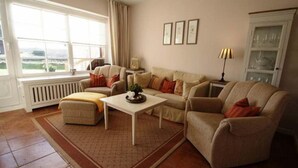 A living room with a sofa set, a coffee table, and a radiator. There are three framed pictures on the wall and a lamp on a side table. - Bredland 11 - Whg. 4 - Holiday apartment by the sea in Nieblum: Terrace, garden & 200 (Nieblum)
