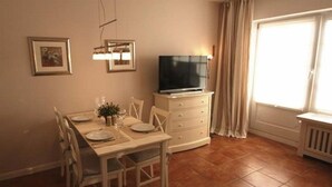 A dining area with a table set for four, a television on a stand, and a window with curtains. - Bredland 11 - Whg. 4 - Holiday apartment by the sea in Nieblum: Terrace, garden & 200 (Nieblum)