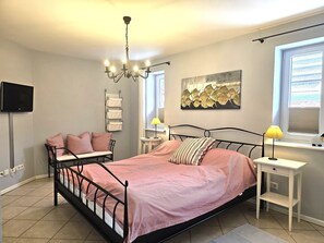 Un dormitorio con cama, mesitas de noche, televisor, candelabro, ventana con persianas y estante montado en la pared.