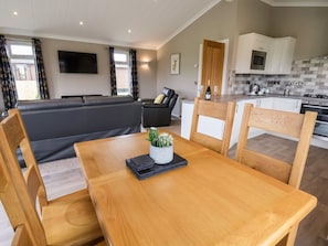 A modern living room with a sofa, a dining table with chairs, a kitchen area with cabinets and appliances, and a flat-screen TV mounted on the wall.