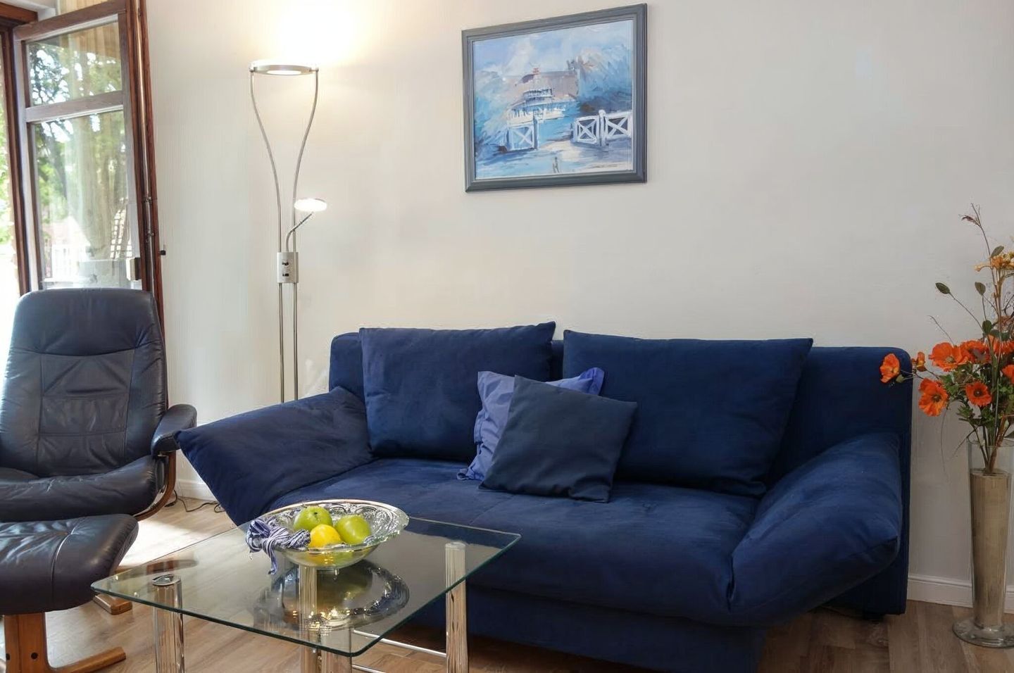 Una sala de estar con un sofá azul, una mesa de centro de cristal con fruta, una silla negra, una lámpara de pie y un cuadro en la pared.