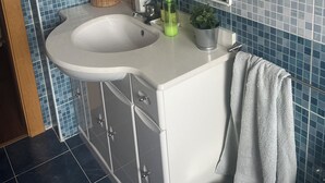 Un baño con lavamanos blanco, un espejo, toallero y repisa con artículos de aseo.