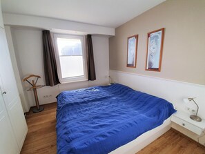 Ein Schlafzimmer mit einem Bett, einem Fenster mit Vorhängen, einem Nachttisch mit Lampe und zwei gerahmten Bildern an der Wand.