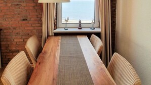 Una habitación con una mesa de madera, sillas a rayas, una lámpara colgante, un muro de ladrillo y una ventana con vista al mar.