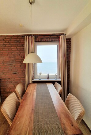 Ein Zimmer mit Holztisch, gestreifter Sitzgelegenheit, einer Hängelampe, einer Backsteinwand und einem Fenster mit Meerblick.