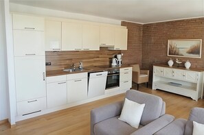 Eine moderne Küche mit weißen Schränken, einer Holzauskleidung und einer Ziegelwand. Es gibt eine Essecke mit einem grauen Sofa und einem weißen Couchtisch.