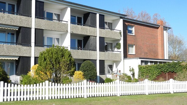 Un edificio residencial de varios pisos con balcones, una cerca de piquetes blancos y una sección de ladrillo.