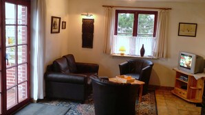 Ein Wohnzimmer mit einer Couch, Sesseln, Hocker, Fernseher und einem Fenster mit Vorhängen.