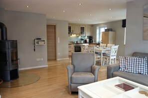 A modern living room with a fireplace, a dining area, and a kitchen in the background. - Golfoase 2 - Semi-detached house for 6 people - Semi-detached house Golfoase 2 for 6 (Oevenum)