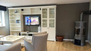 Una sala de estar moderna con televisor, armarios blancos, un sofá gris y una estufa de leña.