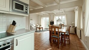 A kitchen with white cabinets, a microwave, a knife block, and a dining area with wooden chairs and a table. - Triibergem 39 - DHH - Holiday home in Utersum: 75 m², garden, 500 m to the beach, (Utersum)