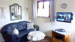 Ein Wohnzimmer mit einem dunkelblauen Sofa, einem kleinen runden Tisch mit einer blumigen Tischdecke, einer Holzkommode, einem Fernseher und einer Wanduhr.